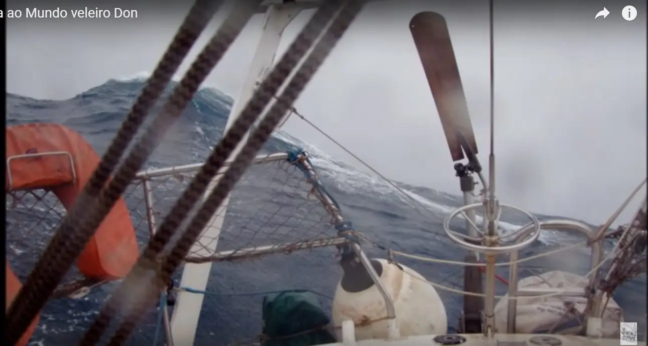 Solo sailor on a sailboat in heavy weather with self-steering windvane visible on the transom